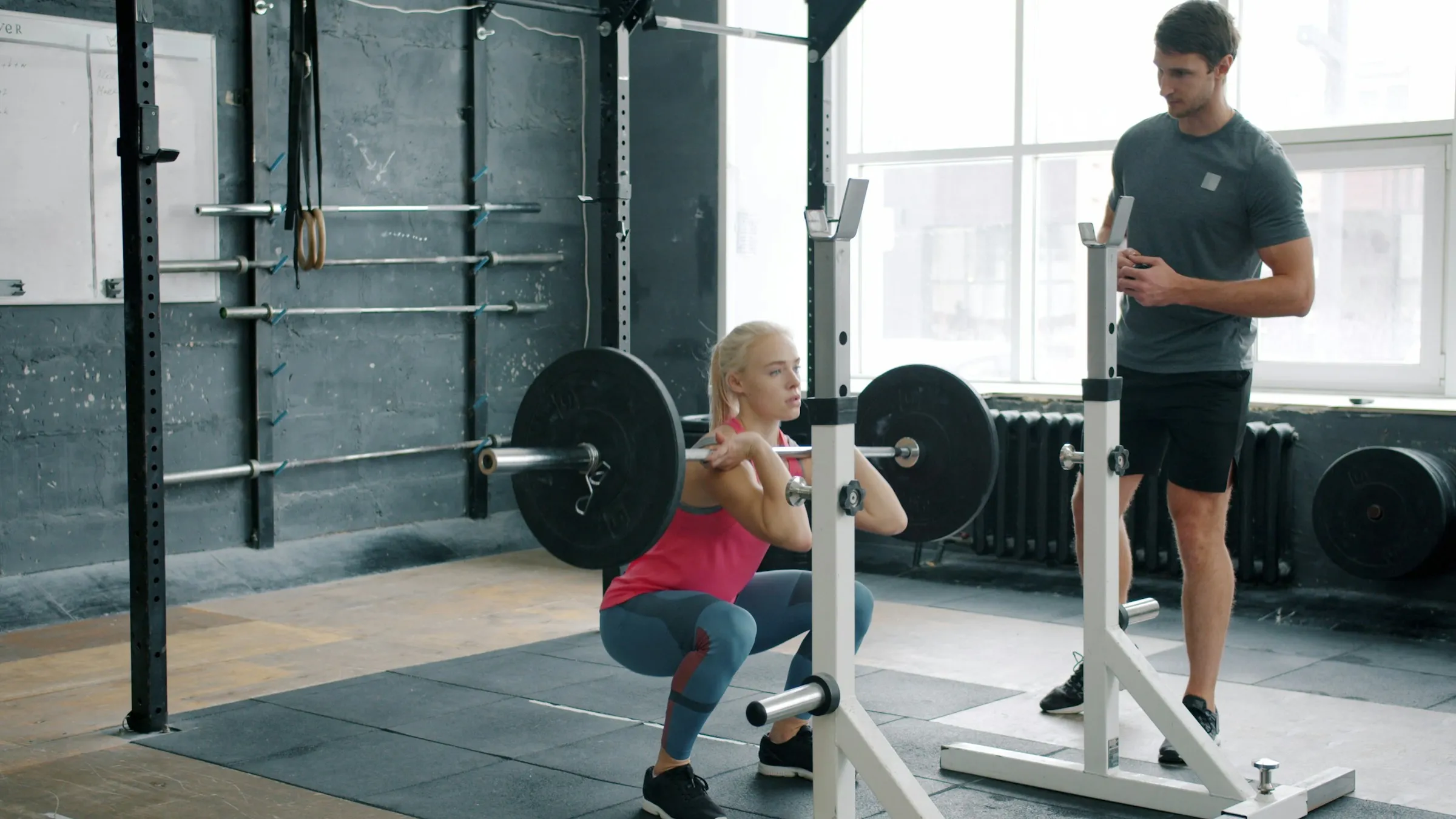 A lifter at the bottom of a squat with a loaded barbell across her shoulders in a gym rack, while a second person stands alongside watching the set