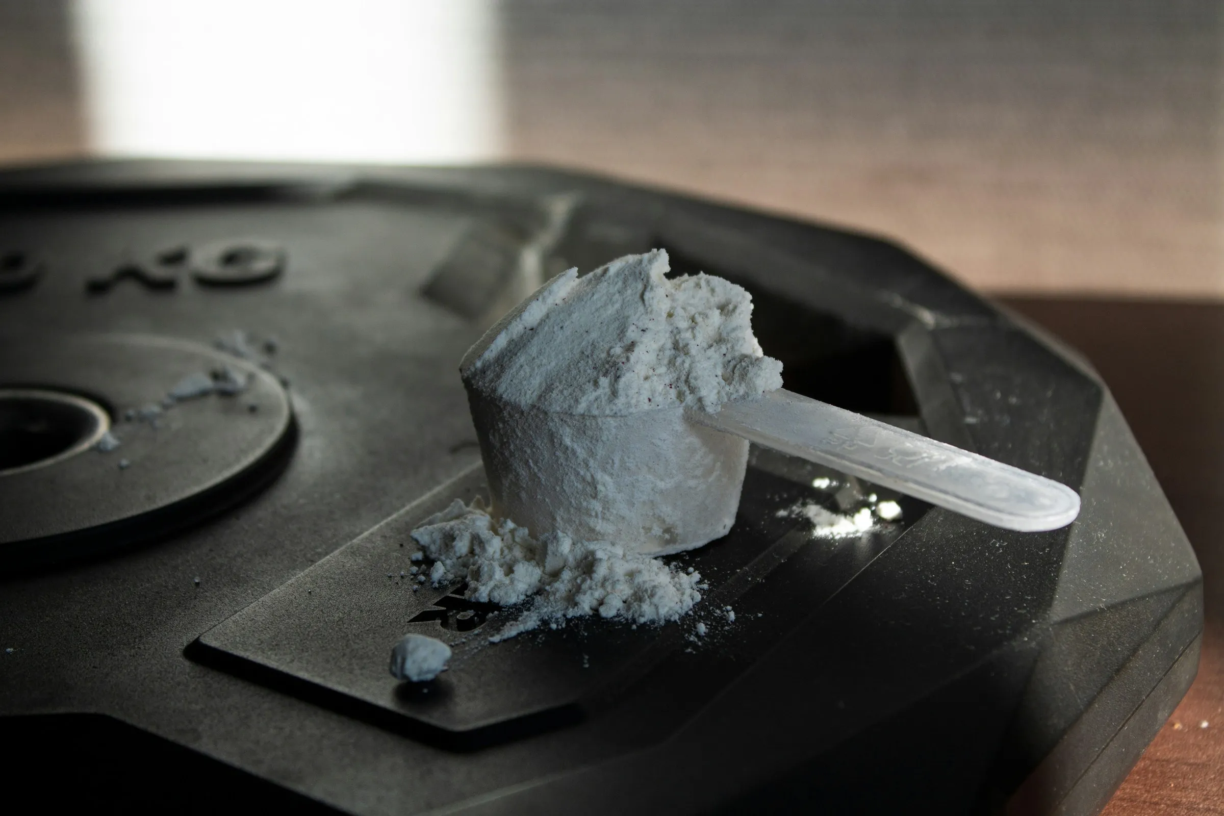 A plastic scoop heaped with white supplement powder resting on a black rubber weight plate, with loose powder spilled around it